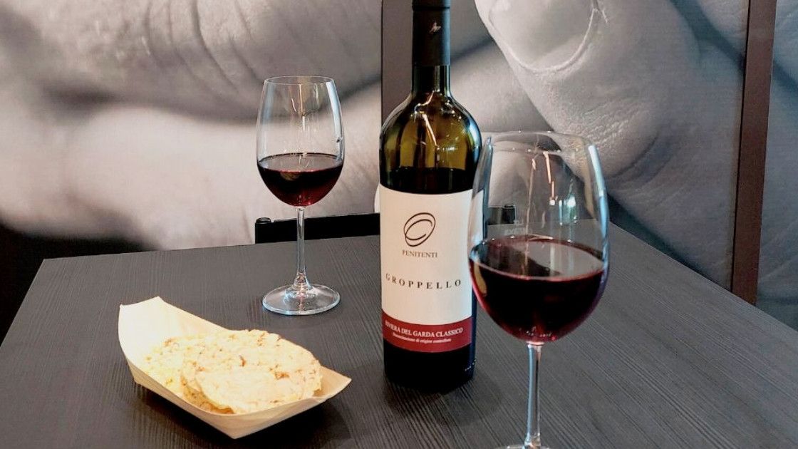Close-up of a bottle of Penitenti Groppello Riviera del Garda Classico red wine on a table with two filled wine glasses and a small wooden tray of crackers.