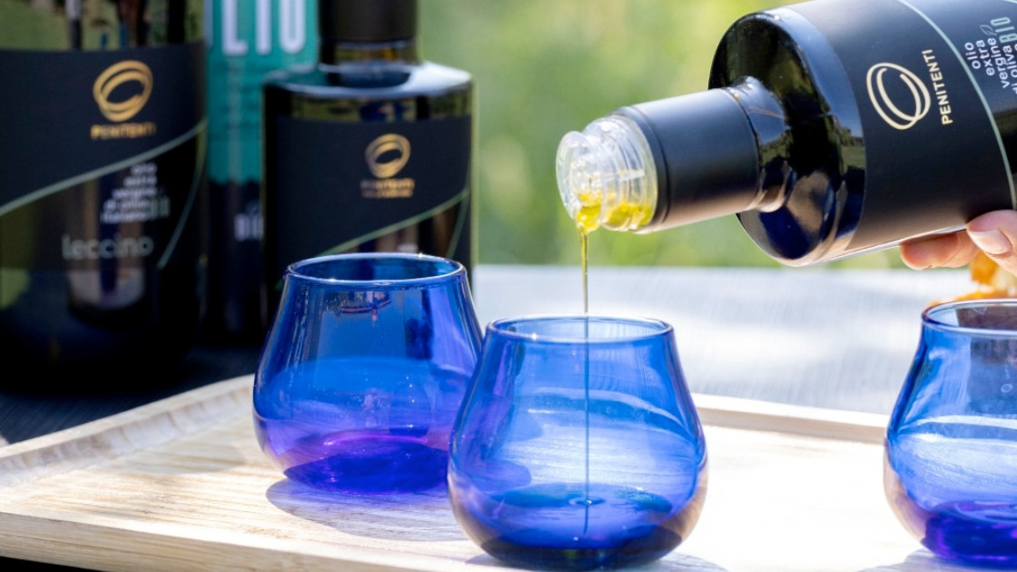 Close-up of Penitenti organic extra virgin olive oil being poured into professional blue glass tasting cups, with dark oil bottles including a Leccino variety in the background.