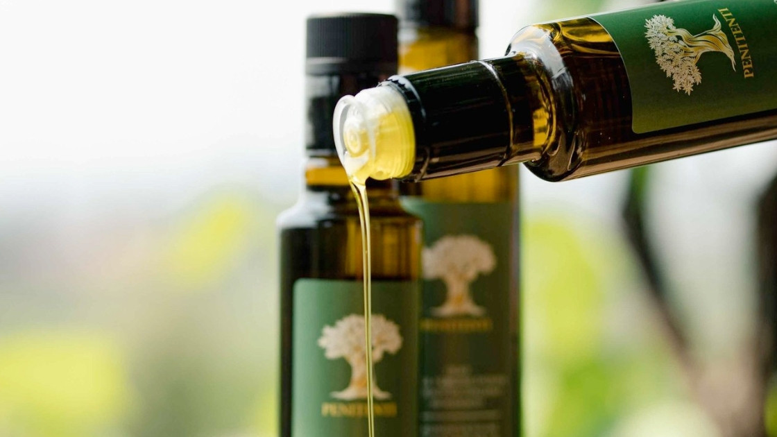 Close-up of golden extra virgin olive oil being poured from a Penitenti bottle with a green label, featuring other oil bottles in a soft-focus background.