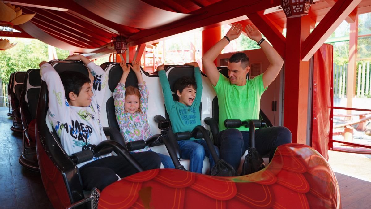 Happy father and three children with raised arms laughing on the Kung Fu Panda Master roller coaster at Gardaland, seated in a colorful themed dragon car.