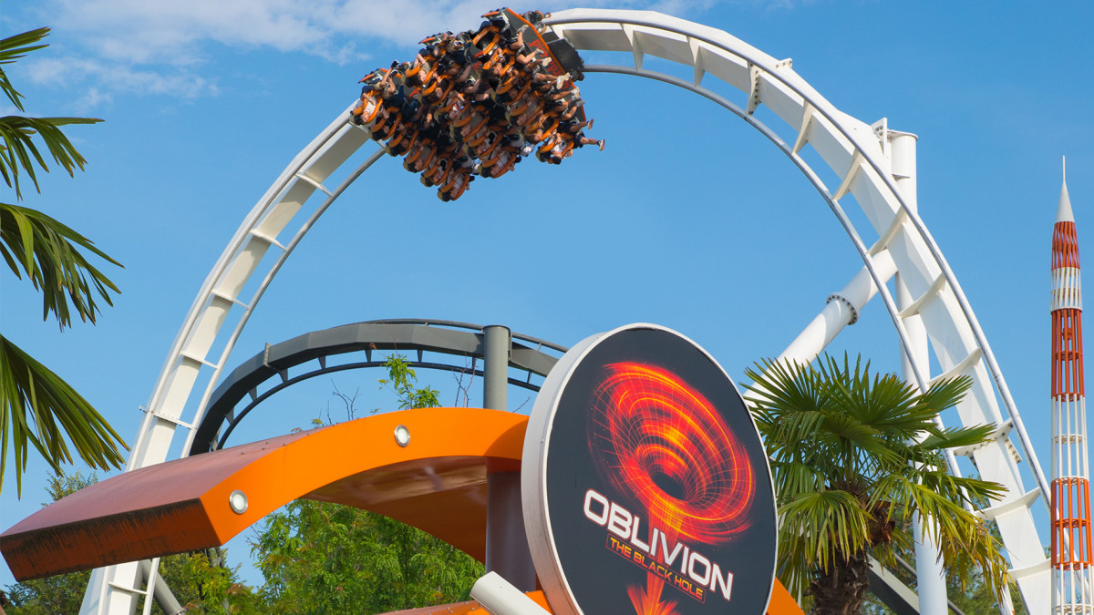 Low-angle view of the Oblivion: The Black Hole dive coaster at Gardaland, showing a vertical drop with passengers suspended over the entrance sign against a bright blue sky.