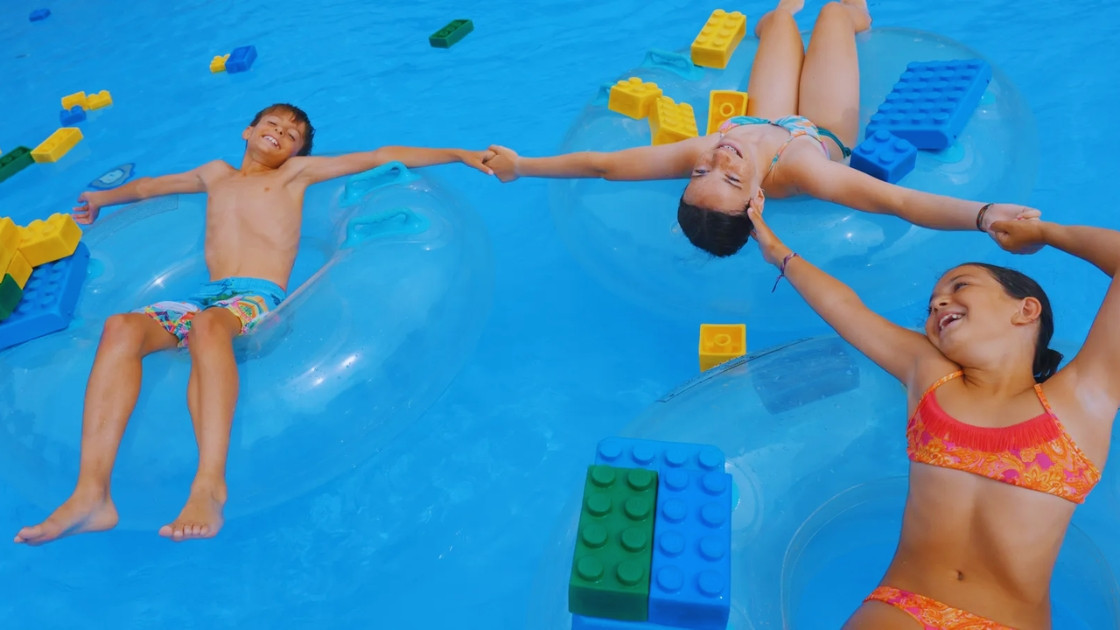High-angle view of three smiling children holding hands while floating on transparent tubes in a blue pool at LEGOLAND Water Park, surrounded by large colorful floating LEGO bricks.