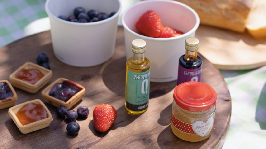 A gourmet picnic spread on a wooden board featuring mini jam tarts, fresh blueberries, strawberries, and small bottles of extra virgin olive oil and balsamic vinegar.