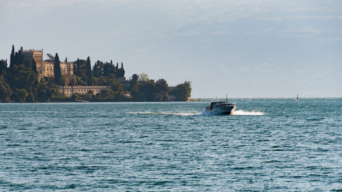 A fast motorboat cruising across the blue waters of Lake Garda, leaving a white wake, with the historic Isola del Garda villa visible in the distance