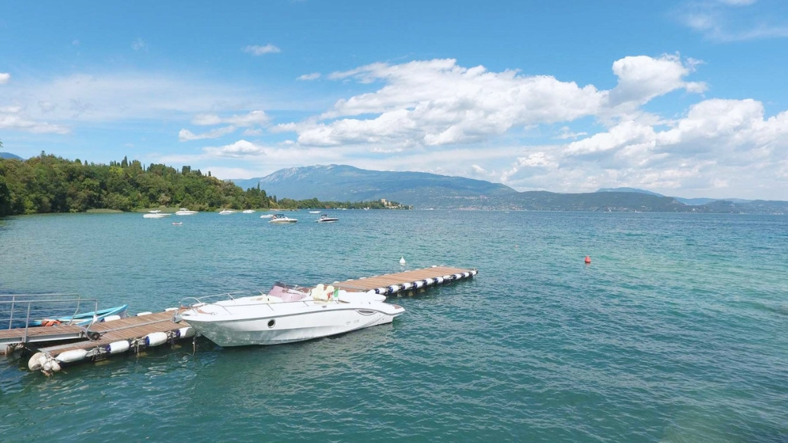 A white motorboat docked at a wooden floating pier on the turquoise waters of Lake Garda, with mountains and a clear blue sky in the background.