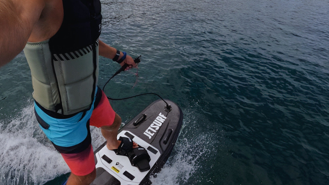 Point-of-view (POV) shot of a rider on a motorized JetSurf board, holding the hand controller and carving through blue water, wearing a safety vest and boardshorts.