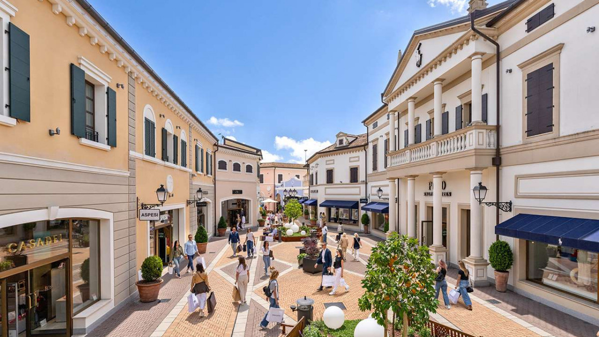 Sunny walkway with shoppers and high-end fashion stores at Noventa di Piave Designer Outlet village.