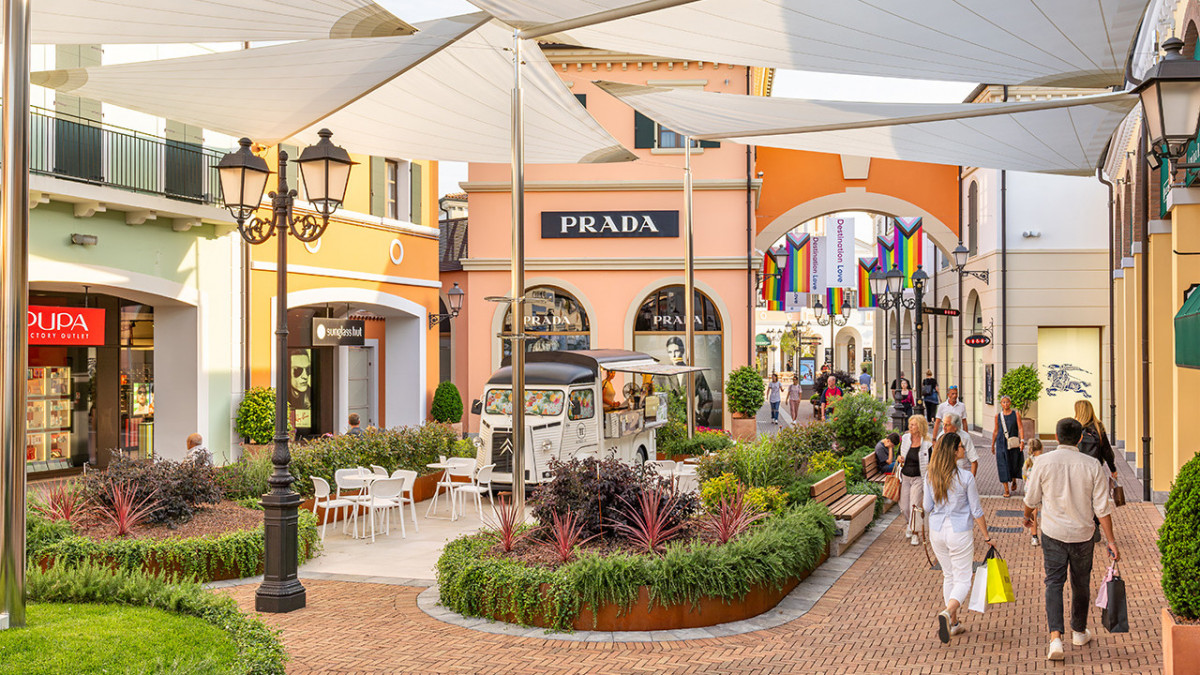 Shoppers walking past Prada and Burberry stores under white sun sails at Noventa di Piave Designer Outlet.