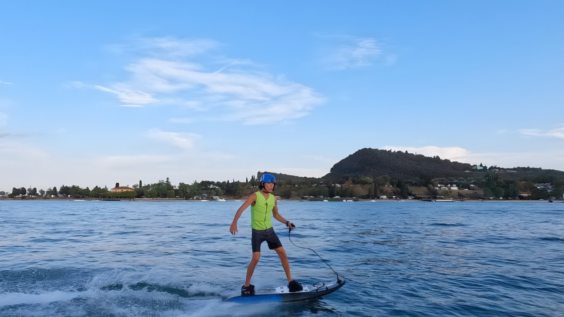 Man wearing a safety helmet and life vest riding a motorized jetboard on Lake Garda with a view of Rocca di Manerba.