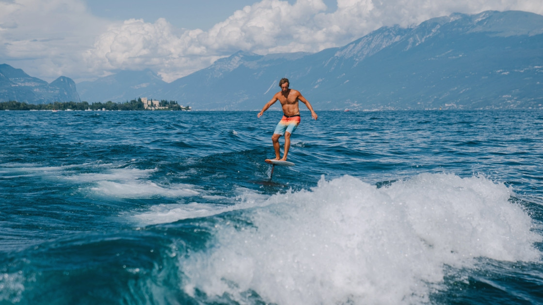 Man surfing a hydrofoil board on Lake Garda with the historic Isola del Garda villa in the background.