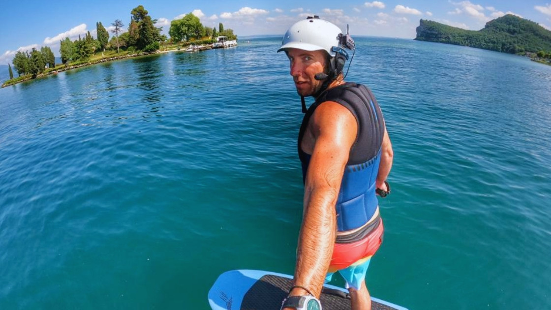 Man riding an electric hydrofoil surfboard on a bright sunny day with a Rocca di Manerba and San Biagio Island (Rabbit Island) on the background.
