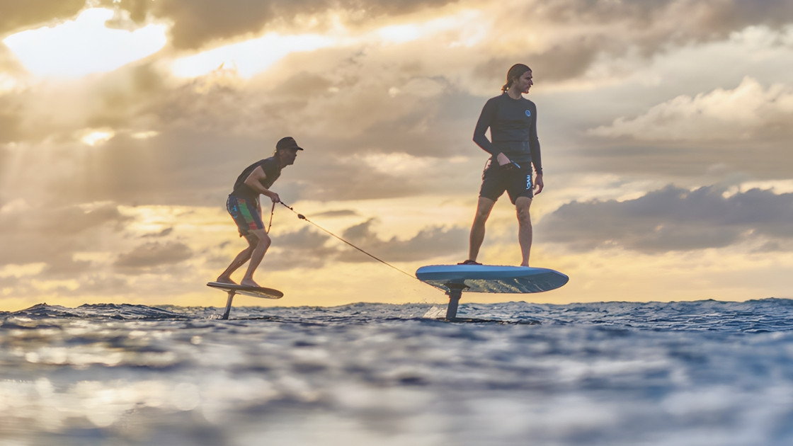 Two men riding e-foils and hydrofoil surfboards on lake waves at sunset with golden sky