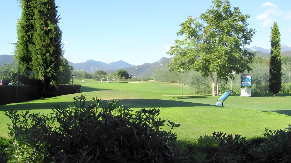 Wide view of a green golf course with mountains in the background, featuring lush trees and a clear blue sky.
