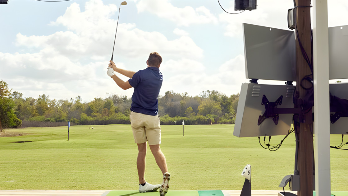 Rear view of a male golfer practicing at an outdoor golf facility with digital tracking monitors and a green fairway background