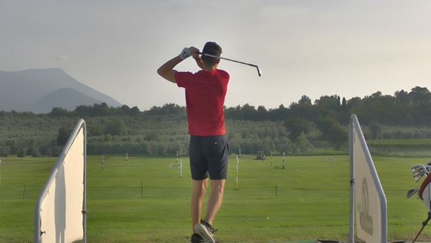 Rear view of a golfer in a red polo shirt finishing a swing on a scenic golf driving range with mountains in the background.
