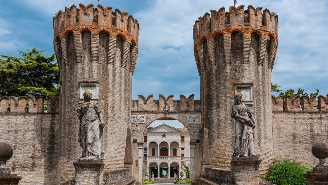 Medieval entrance of Castello di Roncade in Treviso, Italy, featuring two large brick towers, stone statues, and a view of the Renaissance villa through the archway
