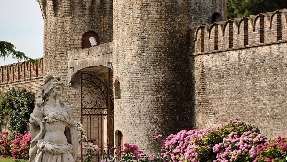 Detail of the medieval brick walls and towers of Castello di Roncade with a historic stone statue and blooming pink flowers in the foreground