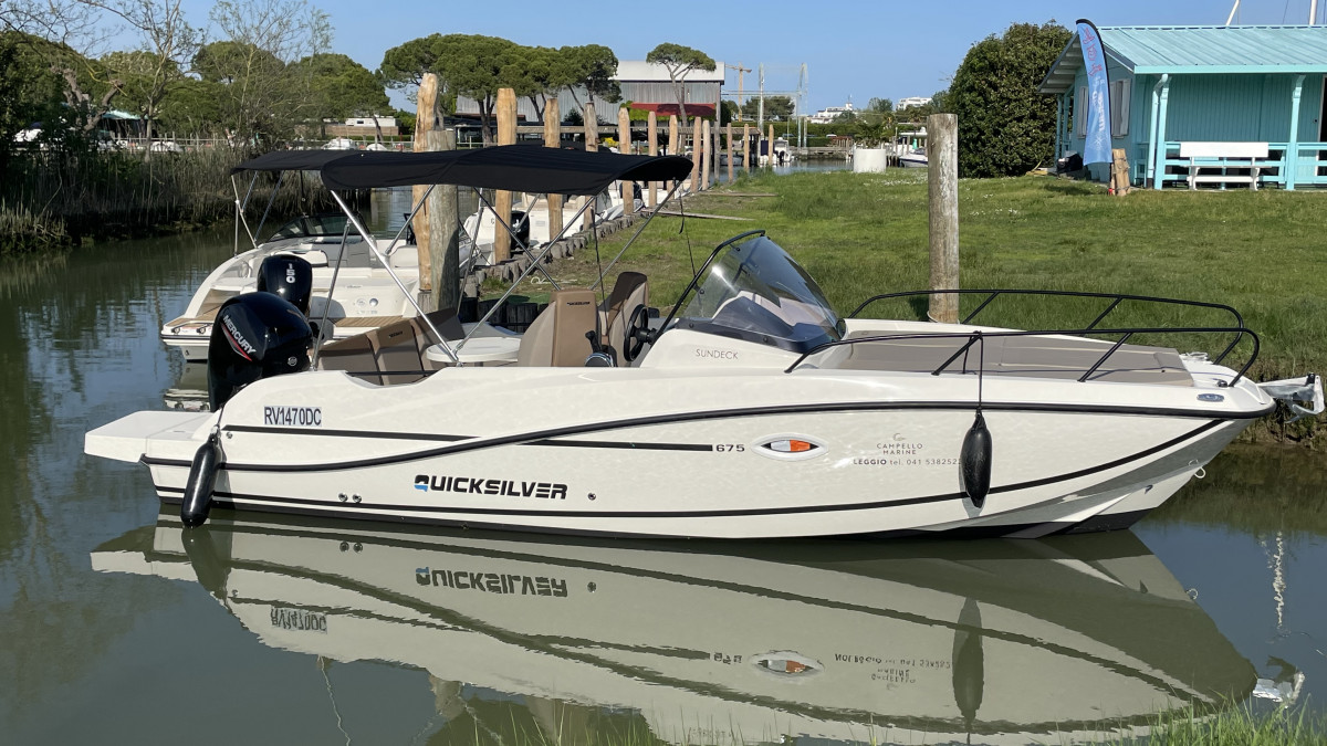 White Quicksilver Activ 675 Sundeck motorboat with a black bimini top and Mercury outboard engine moored in a canal.