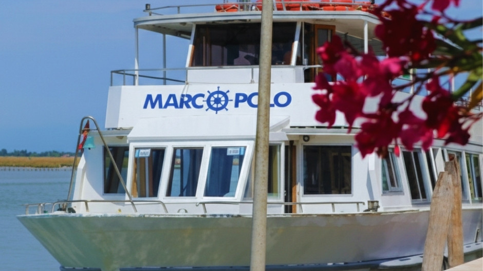 A close-up view of the white multi-deck tour boat 'MARCO POLO', as seen from the side, partially obscured by a central metal post and a prominent spray of deep pink oleander flowers in the right foreground. The boat's blue and white logo, including a ship